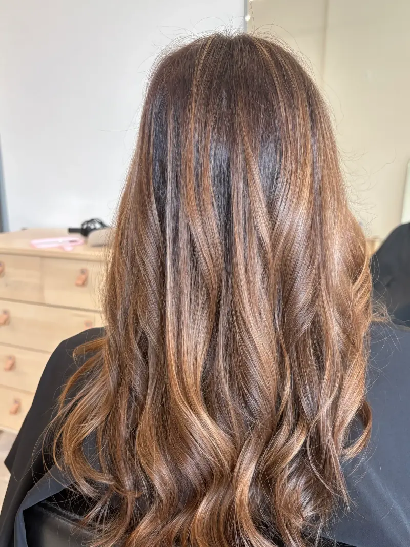 Long brunette hair with caramel highlights, a darker root, and loose waves.